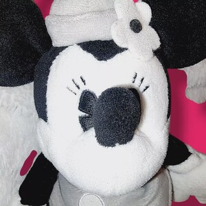Op de afbeelding: Een pluche Minnie Mouse pop met een zwarte neus, zwarte oren en een witte bloem op haar hoofd. De pop is voornamelijk zwart en wit met grijze accenten en een grijze hoed. De achtergrond is felroze.