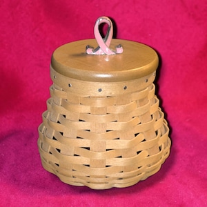 May include: A woven light brown wooden basket with a round, smooth wooden lid. The lid features a silver-toned metal handle shaped like a pink awareness ribbon. The basket has a flared, rounded base and a series of small metal tacks along the rim.