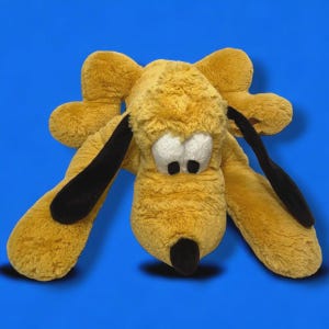 Puede incluir: Un peluche de perro amarillo, con orejas y nariz negras, tumbado sobre un fondo azul. El perro tiene ojos blancos. Este juguete de peluche es un personaje clásico para niños.