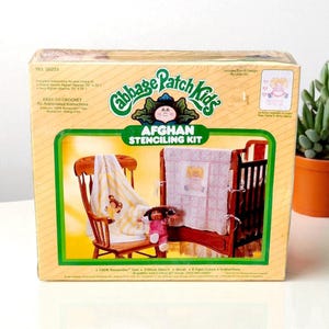 Może przedstawiać: Zestaw do szablonów afgańskich Cabbage Patch Kids w stylu vintage w żółtym pudełku. Pudełko ma zielono-żółty wzór z logo Cabbage Patch Kids. Zestaw zawiera przędzę, szablon, pędzel, kolory farb i instrukcje. Widoczne są drewniane łóżeczko i krzesło.
