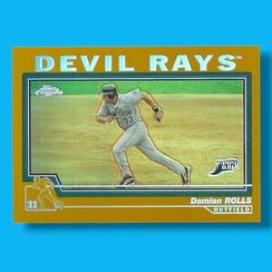 May include: A baseball card featuring Damian Rolls of the Tampa Bay Devil Rays. The card has a gold border with the words "DEVIL RAYS" at the top. The player is in action, running on a baseball field. The card also includes the text "OUTFIELD".
