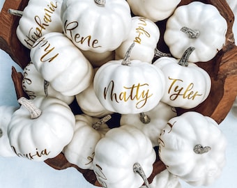 May include: A wooden bowl filled with white pumpkins, each with a gold name written on it. The names include "Rene", "Matty", "Tyler", "Olivia", and "Stephanie".