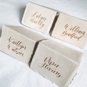 May include: Four place cards with names written in calligraphy on them. The names are Lakyn Hailly, Kaitlyn Watson, Piper Stevens, and William Bradford.