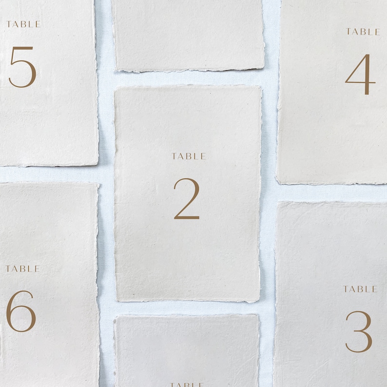 May include: Six white table number cards with brown text. The cards are numbered 2, 3, 4, 5, and 6. Each card has the word "TABLE" above the number.