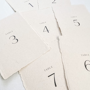 May include: Set of seven table number cards with a rustic, handmade look. Each card is made of textured paper and features a simple black font with the words "TABLE" and the corresponding number.