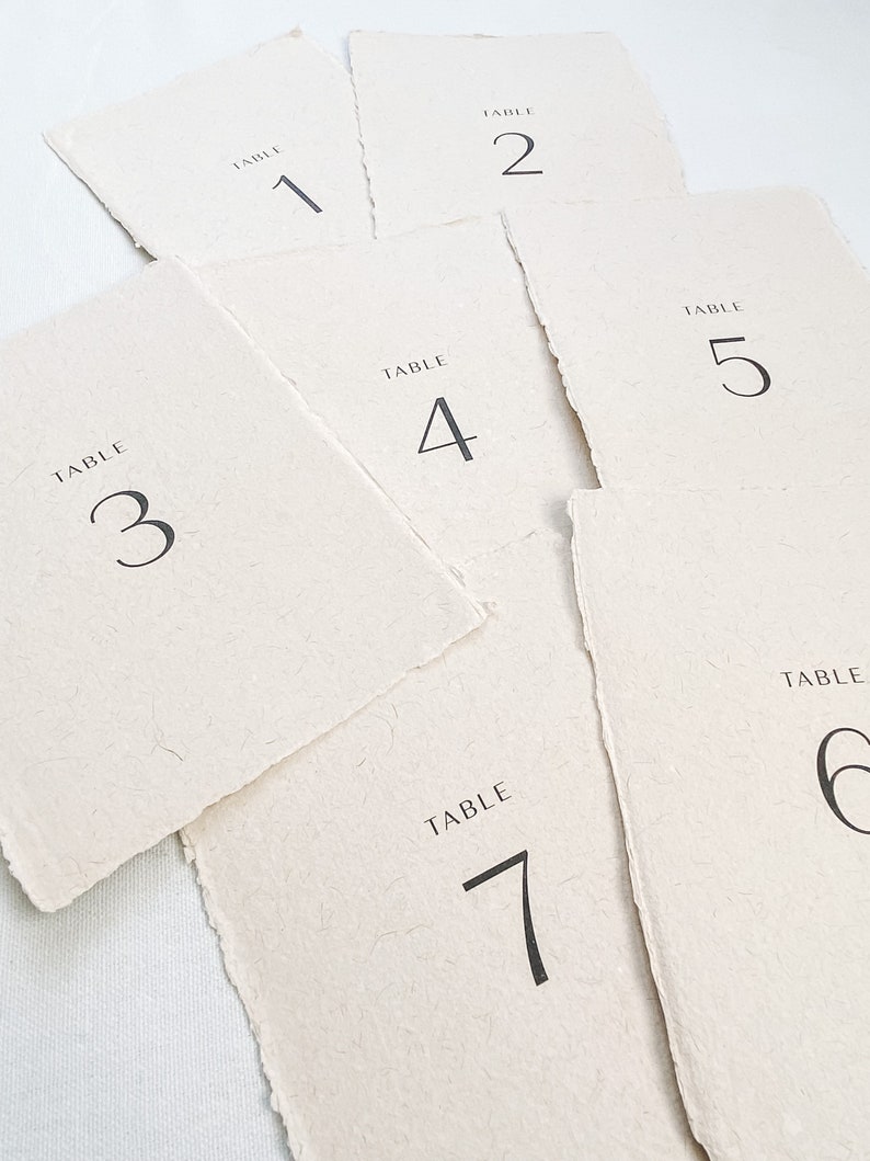 May include: Set of seven table number cards with a rustic, handmade look. Each card is made of textured paper and features a simple black font with the words "TABLE" and the corresponding number.