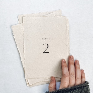 May include: A white, textured paper table number card with the text "TABLE 2" printed in black.