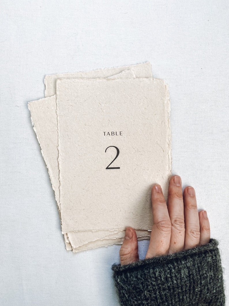 May include: A white, textured paper table number card with the text "TABLE 2" printed in black.