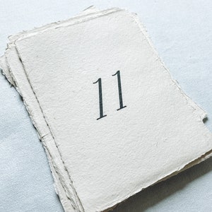 May include: A white paper card with the number 11 printed in black ink. The card has a rough, textured surface and is slightly folded.