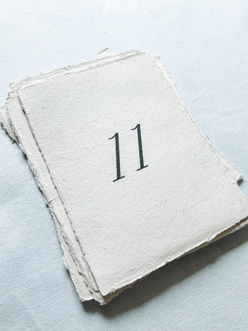 May include: A white paper card with the number 11 printed in black ink. The card has a rough, textured surface and is slightly folded.