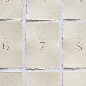 May include: Nine table number cards with a rustic, cream-colored paper texture. The numbers 3 through 11 are printed in a gold font.