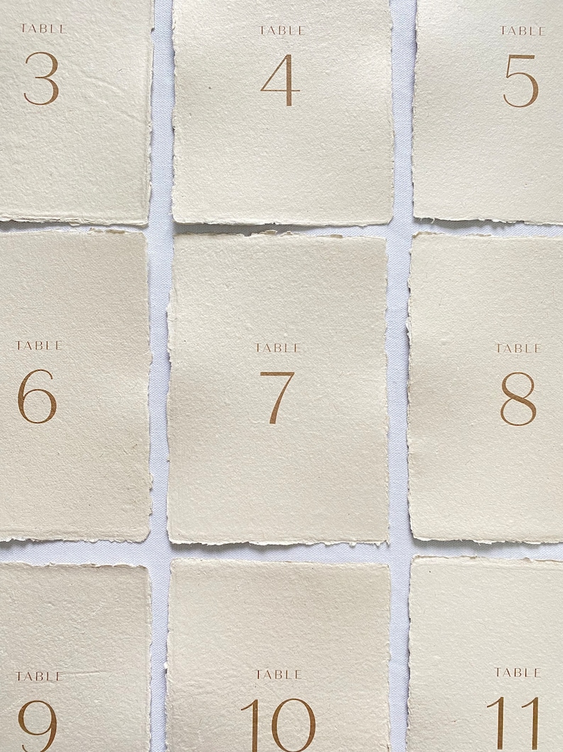 May include: Nine table number cards with a rustic, cream-colored paper texture. The numbers 3 through 11 are printed in a gold font.