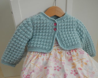 May include: A light blue crocheted cardigan with pink buttons, worn over a white dress with a floral pattern.