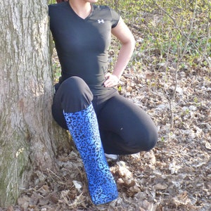 May include: A woman wearing a black shirt with a white logo and black trousers is sitting in a forest. She is wearing a blue and black patterned leg cast.