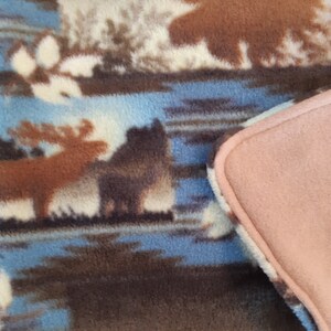 May include: A fleece blanket with a wilderness design, showing moose, trees, and a lake in brown, blue, and cream. The blanket has a soft texture and a pink border.
