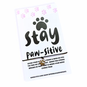 May include: A black string bracelet with a silver paw print charm. The card attached to the bracelet reads "Stay Paw-sitive" and includes instructions on how to make a wish with the bracelet. The card also includes the website address www.etsy.com/shop/sprinklesandwishes.