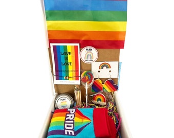 May include: A white gift box filled with rainbow-themed pride items, including socks, a candle, a lollipop, earrings, a sticker, and a card that says "Love is Love".