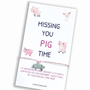May include: Pink and white pig illustrations surround the text "MISSING YOU PIG TIME" on a white card with a pink string bracelet attached. The bracelet has a silver pig charm.