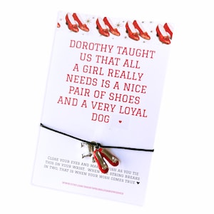 May include: A white card with red text that reads "Dorothy taught us that all a girl really needs is a nice pair of shoes and a very loyal dog." The card has a black string with a charm of red shoes.