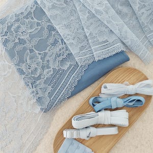 Può includere: Un tessuto in pizzo floreale azzurro stratificato su un tessuto blu a tinta unita, accompagnato da un vassoio in legno contenente quattro rotoli di nastro elastico nei toni del bianco e del blu, insieme a un piccolo set di ganci e occhielli.