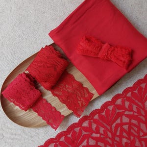 May include: A selection of red fabric and lace. The image shows a folded square of red fabric, several rolls of red lace trim, and a large piece of red lace with a floral design, all arranged on a wooden tray.