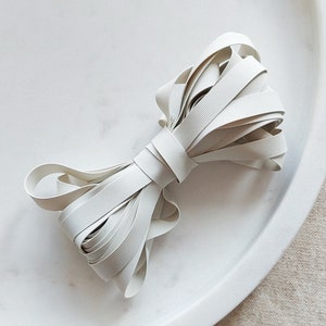 Può includere: Un nastro elastico bianco, piegato e legato a forma di fiocco, è mostrato su una superficie di marmo bianco.