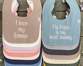 May include: Two stacks of multi-layered muslin baby bibs with snap closures. The left stack features the text I love my Emmie on a tan bib. The right stack features the text My Poppi is my best buddy on a dusty blue bib. Both stacks include various solid colors.