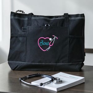 Puede incluir: Bolso tote negro con costuras blancas y m&uacute;ltiples bolsillos. El bolsillo delantero presenta un coraz&oacute;n rosa con un dise&ntilde;o de estetoscopio y el nombre "Joni" y "CNA" bordado en turquesa. Un estetoscopio y un bloc de notas est&aacute;n sobre la mesa.