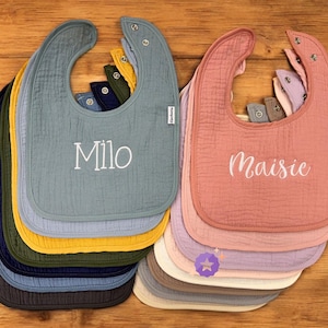 May include: A stack of baby bibs in various colors, including blue, pink, yellow, and green. The bibs are made of a textured fabric and feature snap closures. One bib is embroidered with the name "Milo," and another with "Maisie."