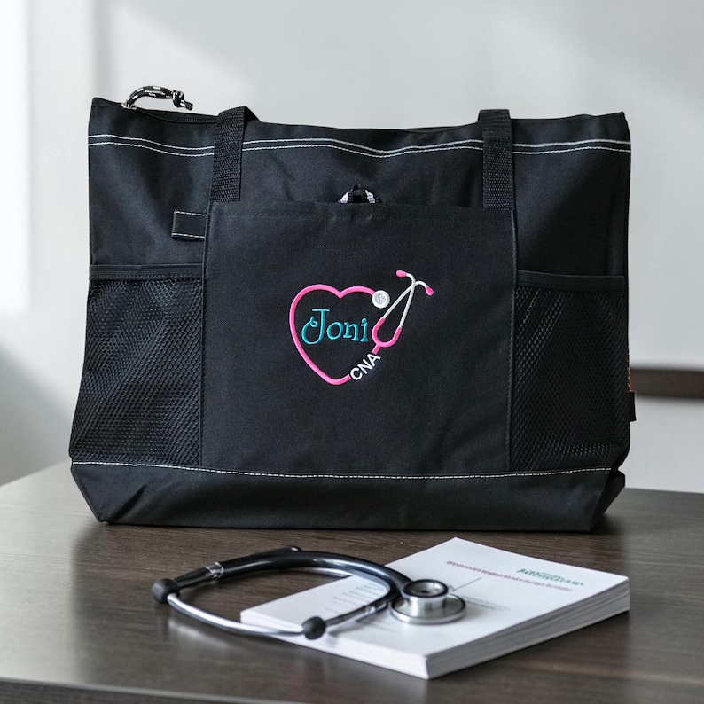 Puede incluir: Bolso tote negro con costuras blancas y m&uacute;ltiples bolsillos. El bolsillo delantero presenta un coraz&oacute;n rosa con un dise&ntilde;o de estetoscopio y el nombre "Joni" y "CNA" bordado en turquesa. Un estetoscopio y un bloc de notas est&aacute;n sobre la mesa.
