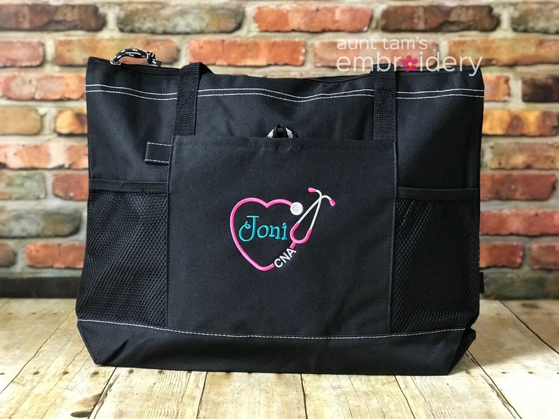 Puede incluir: Bolso tote negro con costuras blancas y m&uacute;ltiples bolsillos. El bolsillo delantero presenta un dise&ntilde;o de estetoscopio en forma de coraz&oacute;n rosa con el nombre "Joni" en turquesa y "CNA" debajo. El bolso tiene asas negras y cremallera blanca.