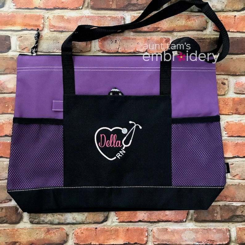 Puede incluir: Un bolso tote morado y negro con una correa negra para el hombro. El bolso tiene un bolsillo frontal con un dise&ntilde;o de estetoscopio en forma de coraz&oacute;n y el nombre "Della RN". Tiene bolsillos laterales de malla y el texto "aunt tam's embroidery".