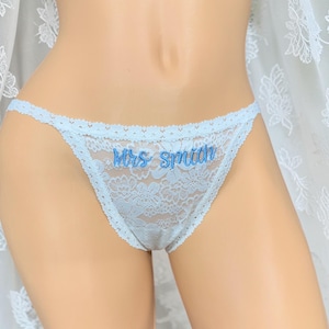 May include: A light blue lace thong featuring a scalloped waistband and leg openings. The front center of the garment is embroidered with the text "Mrs Smith" in a darker blue script font, showcasing a floral pattern throughout the lace fabric.