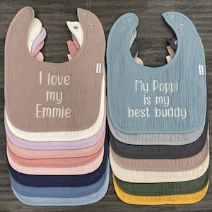 May include: A stack of baby bibs in various colors, including pink, blue, and beige. One bib says "I love my Emmie" and another says "My Poppi is my best buddy" in white embroidery. Each bib has a snap closure.