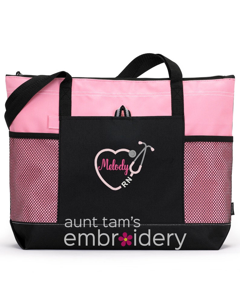 Puede incluir: Bolso tote rosa y negro con cierre de cremallera y asas negras. La parte delantera presenta un bolsillo negro con el texto "Melody RN" dentro de un coraz&oacute;n con un dise&ntilde;o de estetoscopio. La parte inferior de la bolsa tiene el texto "aunt tam's embroidery" con una flor.