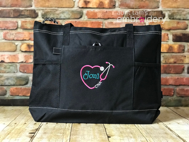 Puede incluir: Bolso tote negro con costuras blancas y bolsillos laterales de malla. El bolsillo delantero presenta un dise&ntilde;o de estetoscopio en forma de coraz&oacute;n rosa con el nombre "Joni" en turquesa y "CNA" en blanco. El bolso tiene asas negras.
