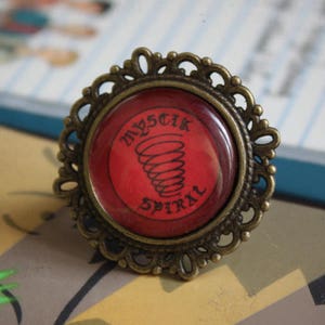 May include: A red and gold ring with a spiral design and the words "Mystic Spiral" on the front.