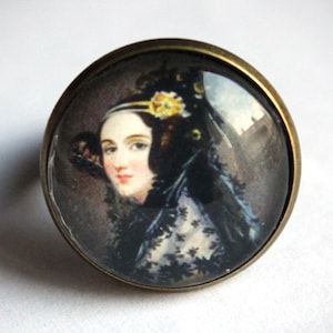 Peut inclure: Une bague en bronze avec un camée en verre représentant le portrait d'une femme vêtue d'une robe en dentelle noire et d'un chapeau à plumes. Le camée est serti dans un bézel rond.