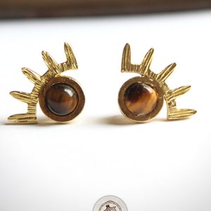 Op de afbeelding: Een paar gouden oorbellen met een tijgeroog-steen in het midden van elke oorbel. De oorbellen zijn gevormd als een gestileerd oog met een waaier van gouden stralen die van boven uitstralen.