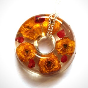 Peut inclure: Un collier avec un pendentif en résine transparente et une chaîne argentée. Le pendentif a la forme d'un beignet et présente des fleurs séchées pressées dans des tons orange et rouge.