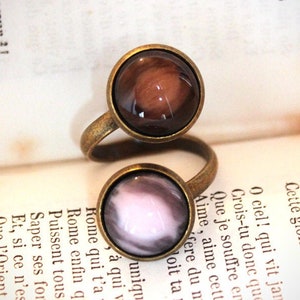Pode incluir: Um anel antigo em tom de latão com duas configurações circulares. A configuração superior apresenta um design marmorizado marrom, enquanto a configuração inferior tem um design marmorizado rosa e branco. O anel está sobre um fundo de papel envelhecido.