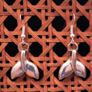 May include: A pair of silver whale tail earrings. The earrings are hanging from a woven wicker background.