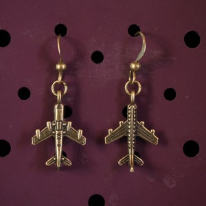 Può includere: Un paio di orecchini a forma di aereo dorati. Gli orecchini sono appesi su uno sfondo viola scuro.