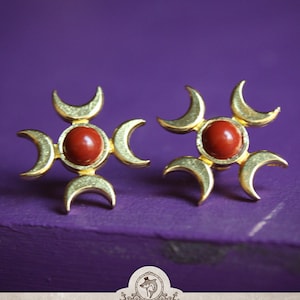 Könnte beinhalten: Ein Paar goldene Ohrringe mit einem roten Stein in der Mitte jedes Ohrrings. Die Ohrringe sind in Form eines vierzackigen Sterns mit Halbmonden für jede Spitze gestaltet.