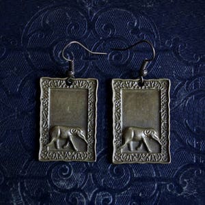 May include: A pair of bronze earrings with a rectangular frame and a detailed design. Each earring features a small, stylized animal figure, possibly an elephant or a tapir, walking to the right.