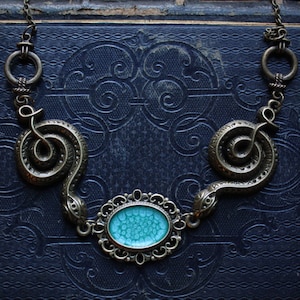 Peut inclure: Un collier en bronze avec deux pendentifs en forme de serpent et une pierre de cabochon bleu turquoise. Le collier est sur un fond bleu foncé.