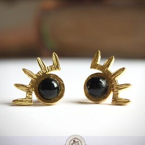 May include: Gold earrings with black gemstone accents in the shape of eyes with lashes.