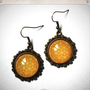 Puede incluir: Un par de pendientes de color dorado con un patrón geométrico redondo de color naranja y blanco. Los pendientes tienen un diseño delicado y adornado.