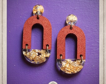 May include: A pair of red and gold earrings with a unique arch design. The earrings are made of resin and feature gold leaf accents.