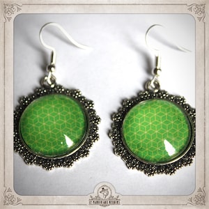 Peut inclure: Une paire de boucles d'oreilles pendantes argentées. Chaque boucle d'oreille présente un motif géométrique vert rond, encadré dans un cadre festonné décoratif. Fermeture à crochet.